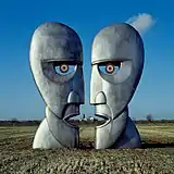 A colour photograph of two large silver-grey iron sculptures of opposing silhouetted faces. The sculptures are standing in a brown wheat field with a blue sky behind them.