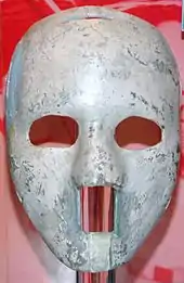 the mask is white and of solid construction with egg-sized oval cutouts for the eyes and a rectangular cutout from the base of the nose to below the lower lip