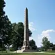 A stone obelisk monument against a blue sky and trees, with the statue of a pioneer in the front.