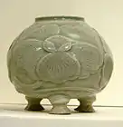 A blue-green tinted grey pot with a wide bottom supported by three stubby legs, a wide body, and a smaller opening at the top. A large five petal flower pattern is out-dented on the body of the jar.