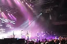 A wide shot of a stage with only four musicians visible: each is playing a type of guitar. A drum kit is mid-stage but the Coghill is obscured by lighting and equipment. The audience are across the front, below stage, some have one fist raised. A photographer is centred, front of stage, another is further to the right with a camera pointing at the band, a third cameraman is at left pointing into the crowd. Behind the group on the left is a large screen which depicts various words including "Fight clouds baby blue despair skin red lies lost stars ith empty rocket paint love g". Overhead lights shine down on the performers. To the right is a large sign with white writing "bring your living room to life" on red-orange background.