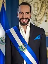 A standing man (Nayib Bukele), wearing a suit and a presidential sash, facing the viewer.