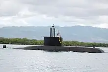 A submarine travelling on the water's surface near to shore, with trees and mountains in the background.