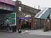 A railway on a brick viaduct crosses a road on a steel bridge, with an entrance below a blue sign reading "SHEPHERD'S BUSH MARKET STATION" in white letters