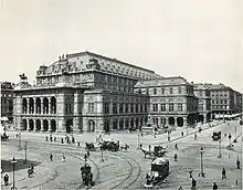An imposing, heavily ornamented building in a city location, with numerous horsedrawn vehicles and pedestrians passing. There are visible tramlines in the street.