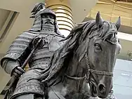 Statue of a man on horseback in full ceremonial armour and holding a mace, in front of a large pillared building.