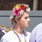 Man seen in profile wearing makeup, with flowers arranged on a headband high on his head; a rainbow-lolored lanyard around his neck reads &quot;Suffolk Pride&quot;