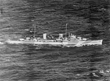 Black-and-white aerial photograph of a two-funnelled cruiser underway. Alternating light and dark bands are painted on the side of the ship.