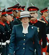 Thatcher in a blue suit and hat, walking in front of troops
