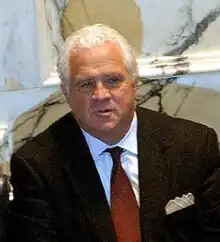 A well-dressed man with white hair sitting before a marble-panelled wall