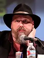 A man in a beard and wide-brimmed hat addresses a crowd at a convention.