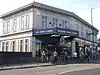 A white-bricked building with a rectangular, dark blue sign reading "WILLESDEN GREEN STATION" in white letters all under a light blue sky