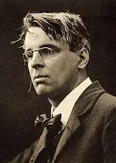 A posed black and white photograph of Yeats. He is wearing smart clothes and spectacles, while his hair looks a bit tousled