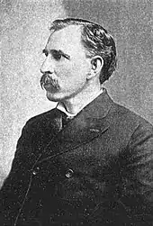 A man in his late forties with black hair and a mustache. He is wearing a tightly-buttoned black coat and facing left.