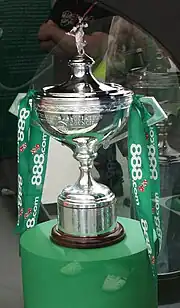 A silver cup-shaped trophy with a trumpet base, a Greek shepherdess finial on top, and two deco square-section handles at the sides with green sponsor ribbons tied to them; the trophy is sitting on a green cylindrical plinth